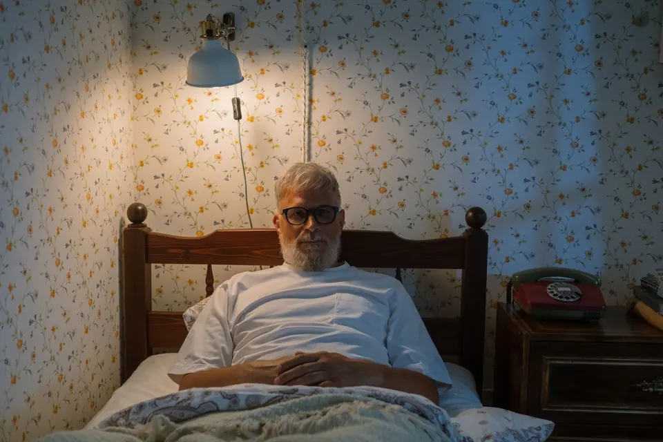 Un homme âgé assis dans son lit, le soir, sous une applique allumée
