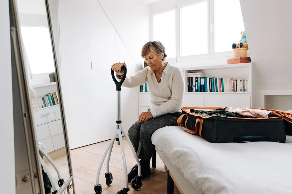 Une femme âgée assise sur son lit, une aide à la marche à portée de main et un sac de voyage ouvert